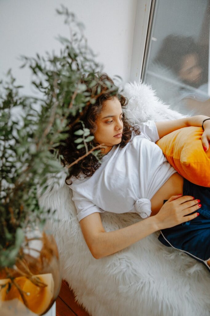 15 Thoughts Women Have On Their periods - stock photo from pexels of a woman laying down by a window with brown hair, a white t-shirt and blue shorts by Polina Zimmerman