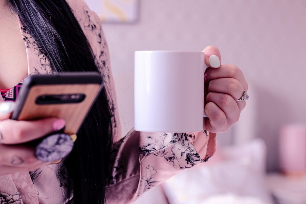 A close up photo of Jordanne holding a white mug in her left hand and a phone in her right. She has on a pink silk robe with black and white flower design.