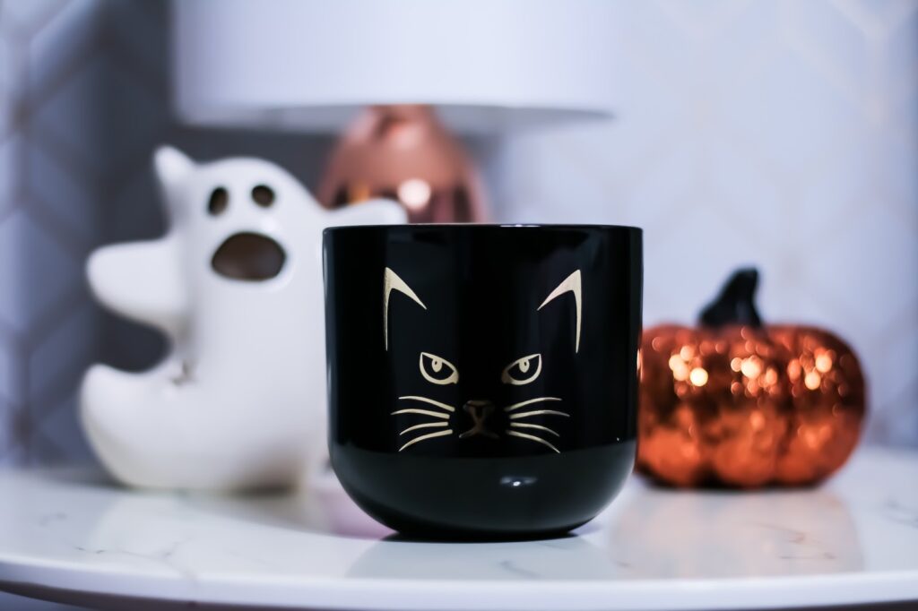 Cat Candle photo of a candle with a cat face on it. Black candle with white ghost candle holder in the background and a bright orange sparkly pumpkin.
