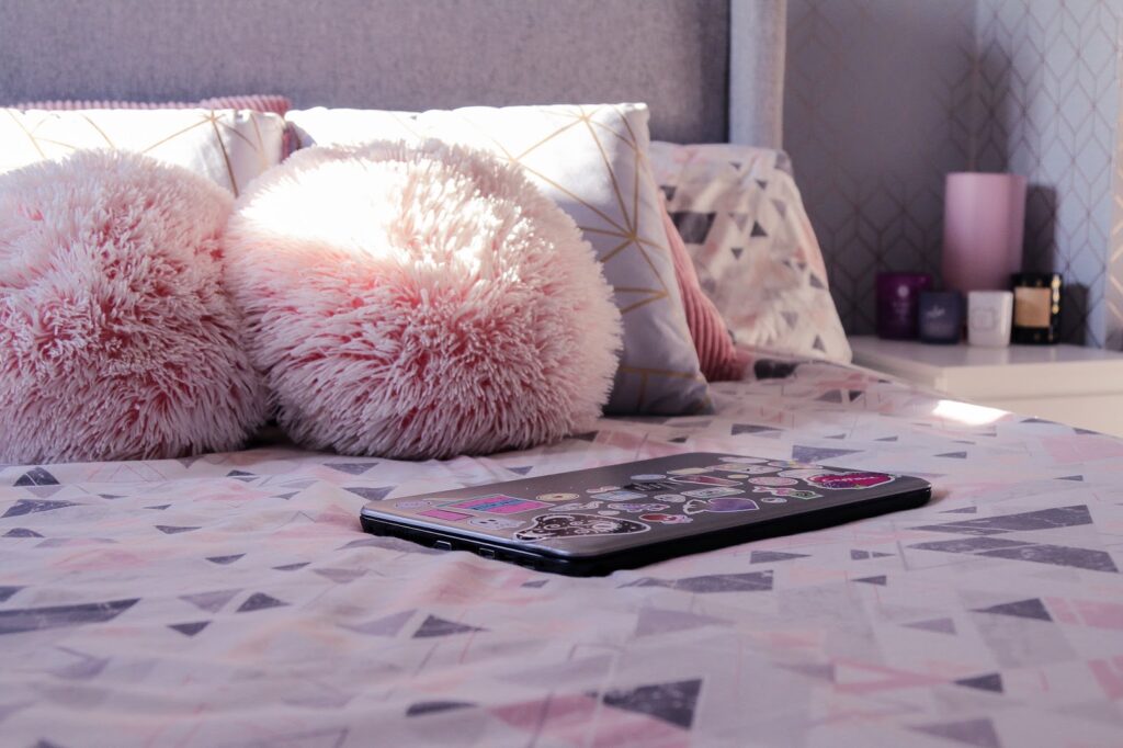 Let's catch up blog post on Thelifeofaglasgowgirl.co.uk Close up of a double bed with a large grey headboard with pink geometric pattern bed sheets. There are 2 large pink pillows, 2 medium great pillows with gold geometric pattern and 2 small round pink fluffy pillows and a silver laptop on the bed and a side table in the background blurred.