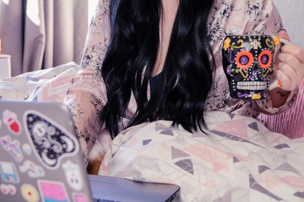 Let's catch up blog post on Thelifeofaglasgowgirl.co.uk Mid shot of a girl with long black hair sitting on a bed with pink geometric bed sheets, wearing a pink silk robe holding a black skull mug with a silver laptop on the bed.