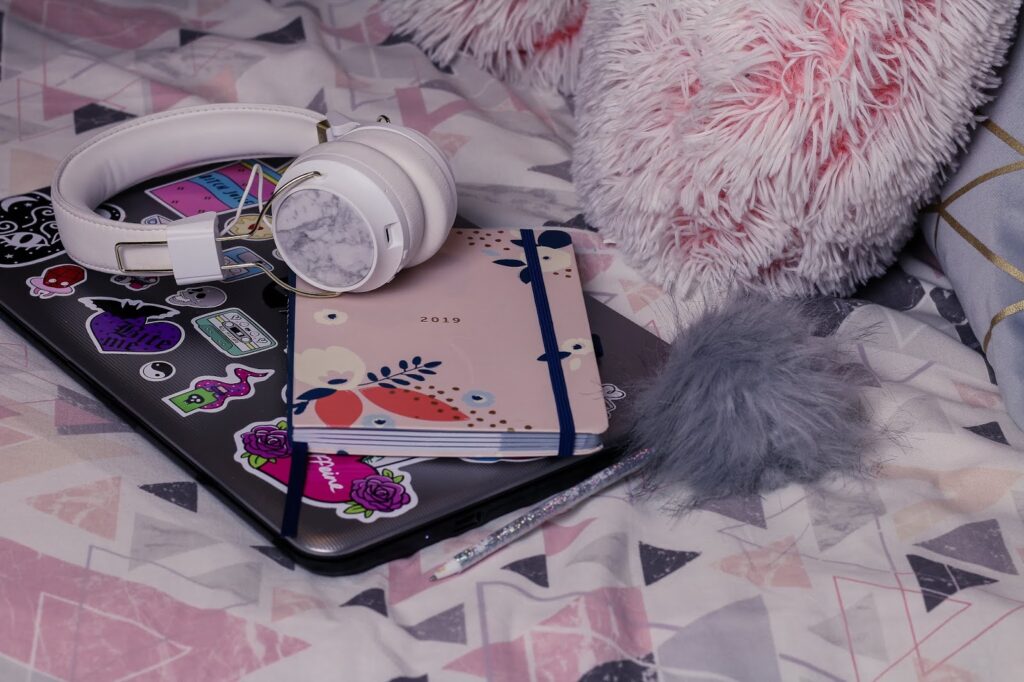 November to do list blog post photo on thelifeofaglasgowgirl.co.uk Close up photo of white headphones, a silver HP laptop and busy b diary that is pink sitting on a bed with geometric pink and great bed sheets.