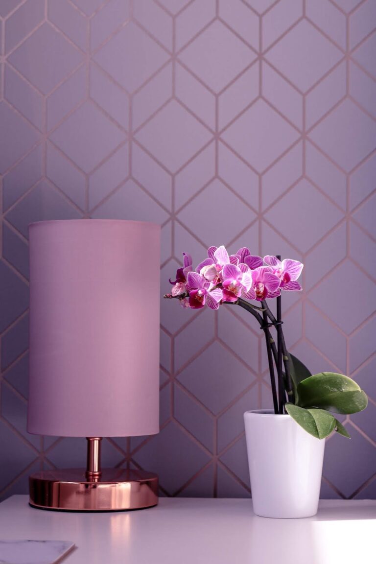 Close up photo of a purple orchid on a white bedside table next to a pink lamp
