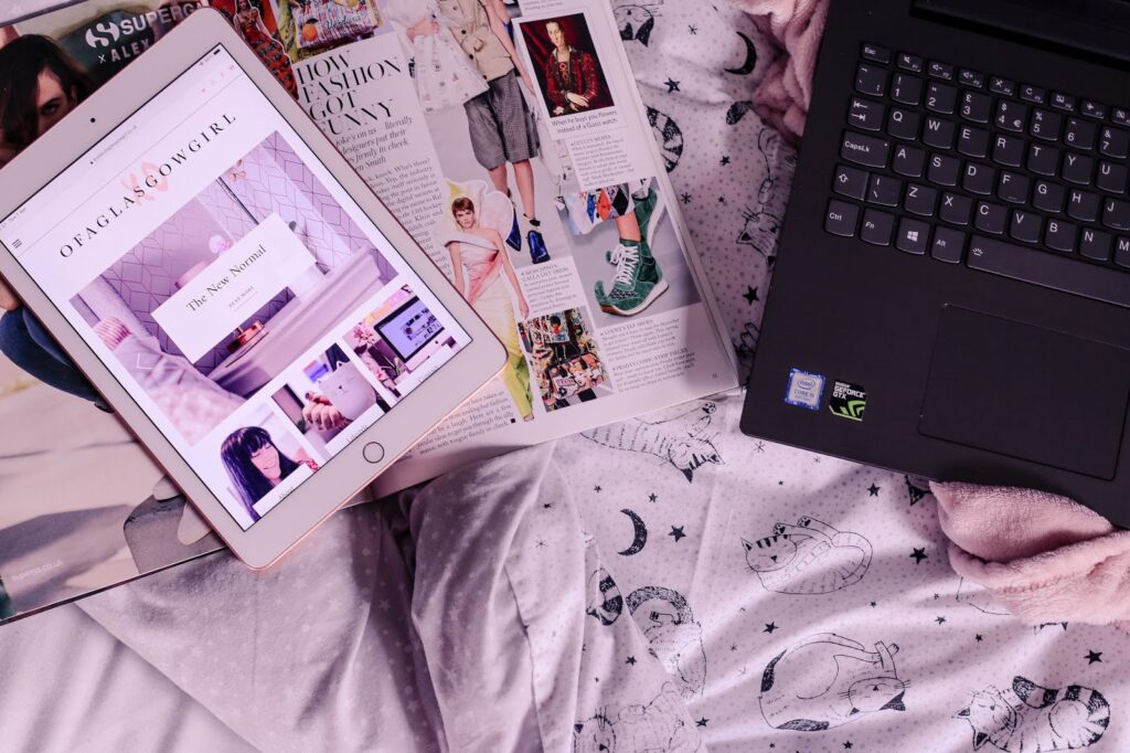 Birds eye view of a white iPad with a vogue magazine under it on top of a bed with a black laptop next to it.