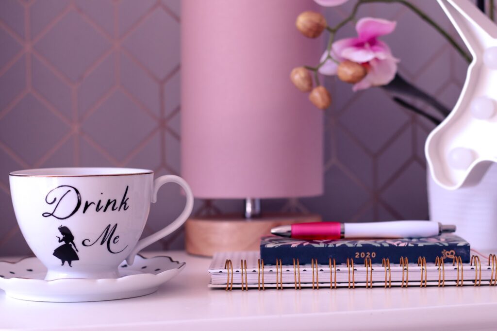 Close up photo of a 2020 diary and a white tea cup on top on a white side table with a pink lamp and faux pink orchid in the background blurred.