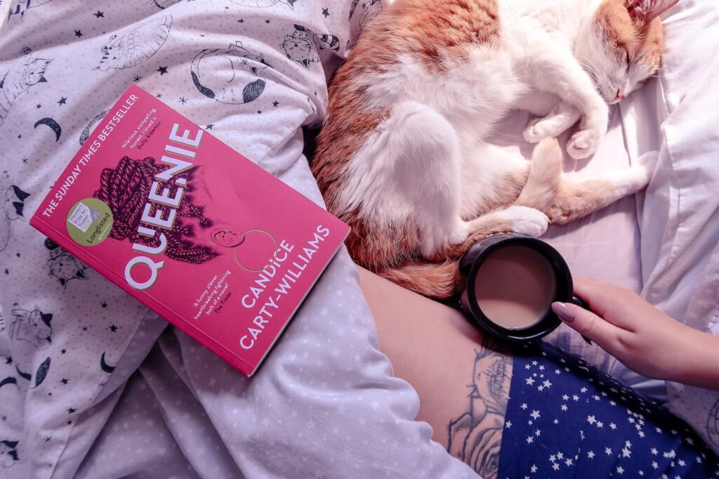Birds eye view of Queenie by Candice Carty-Williams book, pink cover, on a bedsheet that is white with black cat outlines on it.