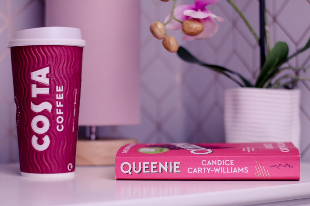 Close up photo of the book Queenie by Candice Carty-Williams on a white bedside table next to a Costa take away cup