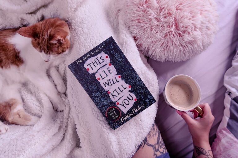 Birds eye view of the book This Lie Will Kill You by Chelsea Pitcher on a white blanket and a cat in the background and pink pillow out of focus with ofaglasgowgirl holding a mug