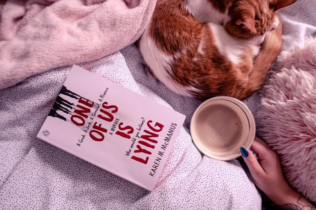 Birds eye view of One of us is lying book on a bed with spotted bedding and a cat to the right with a cup of coffee next to it