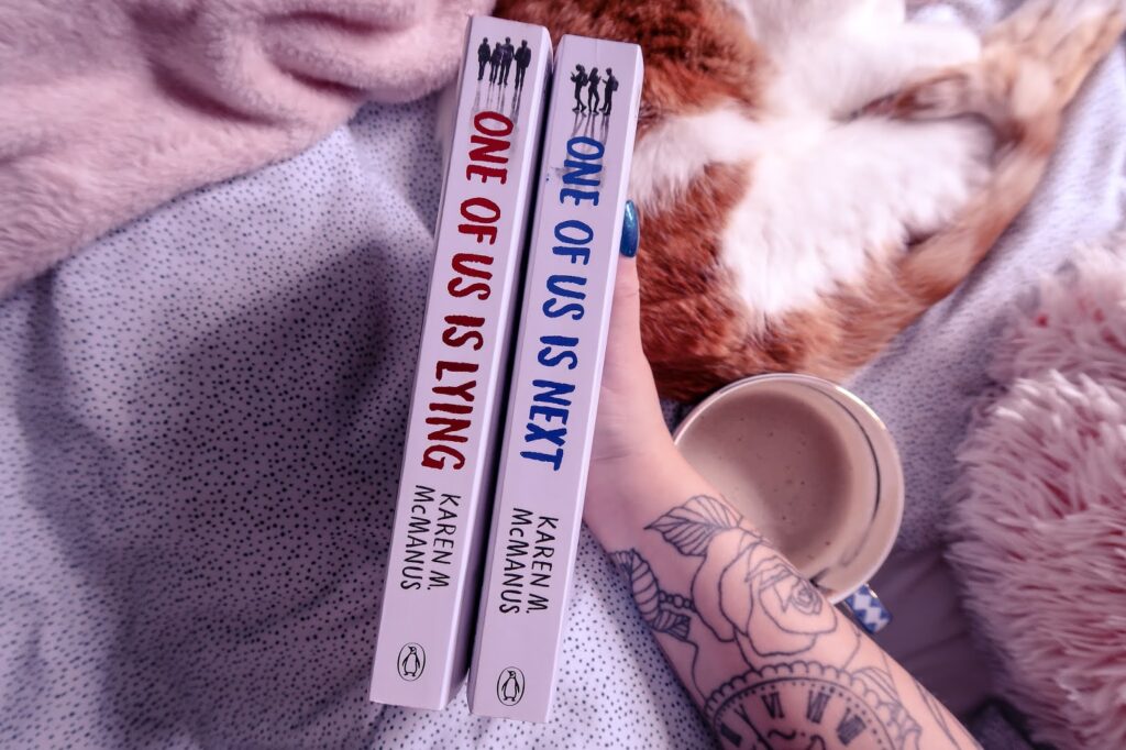 Birds eye view of One of us is lying book and one of us is next book on a bed with spotted bedding and a cat to the right with a cup of coffee next to it