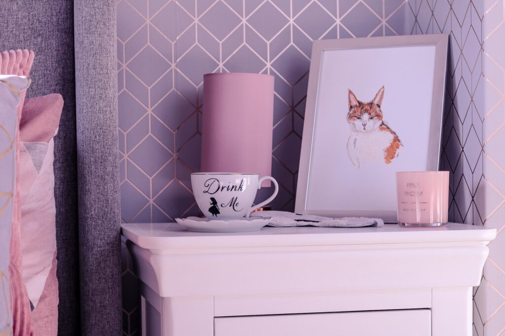 Close up photo of a white mug that has drink me written on it with black ink on top of a white bed side table with a pink lamp and faux pink orchid behind it out of focus.