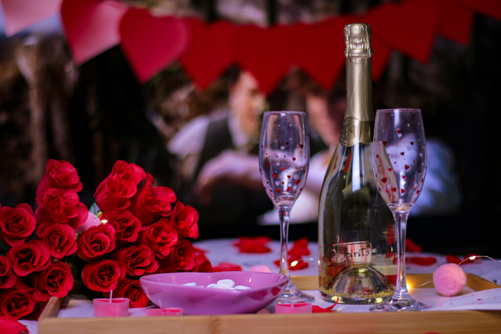 Valentine's date night ideas - Close up photo of a bottle of bubbly and two heart glasses with red roses and candles on a bed