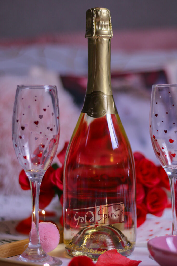 Valentine's date night ideas - Valentine's date night ideas - Close up photo of a bottle of bubbly and two heart glasses with red roses and candles on a bed
