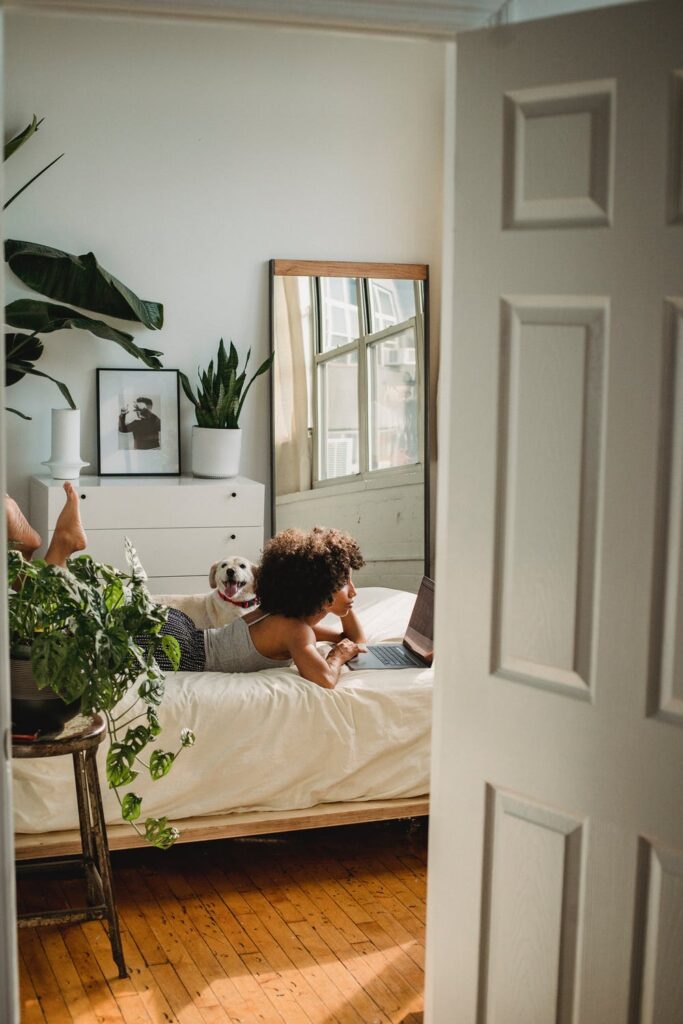 Stock photo of a Black Woman laying on a white bed with a silver laptop and a small dog by Samson Katt on Pexels