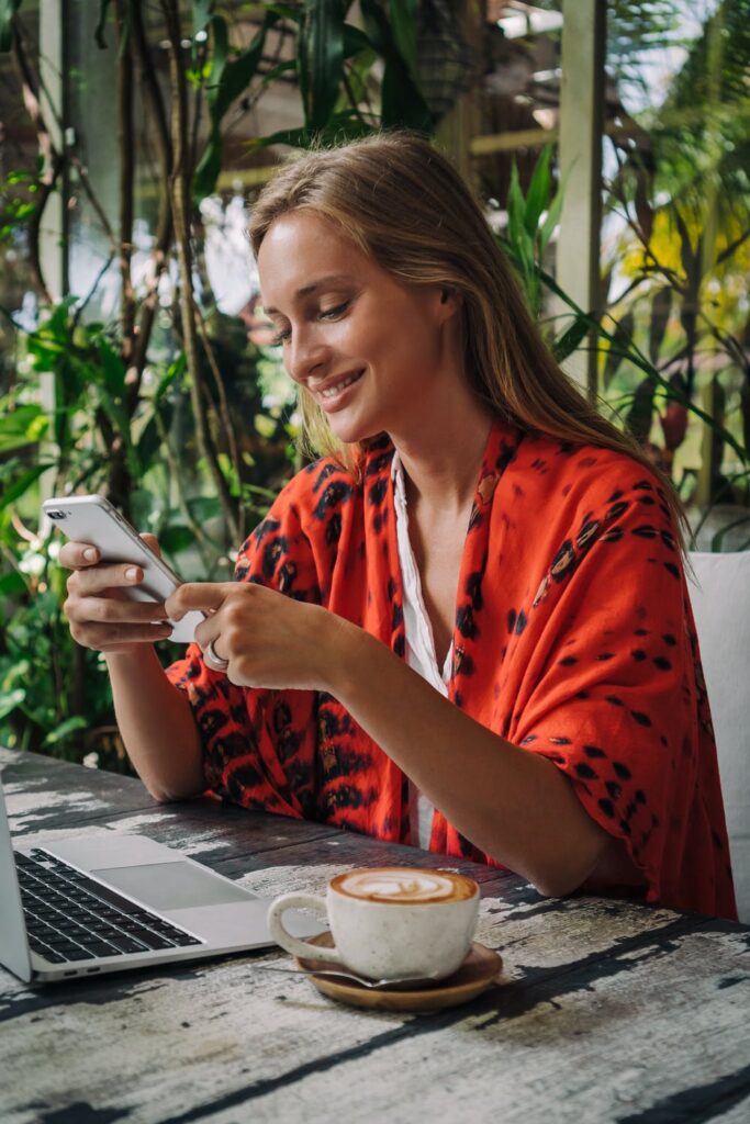Stock photo of a woman sitting at a table with a coffe, phone in hand and smiling wearing a red top by Mikhail Nilov on Pexels