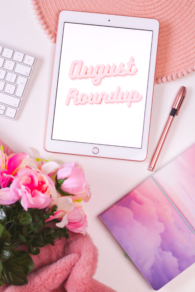 Flatlay photo of an ipad with a graphic that has "August Roundup" written in pale pink on a white background. There are pink flowers in the bottom left corner of the photo and a cloud notebook on the right with a rose gold pen.