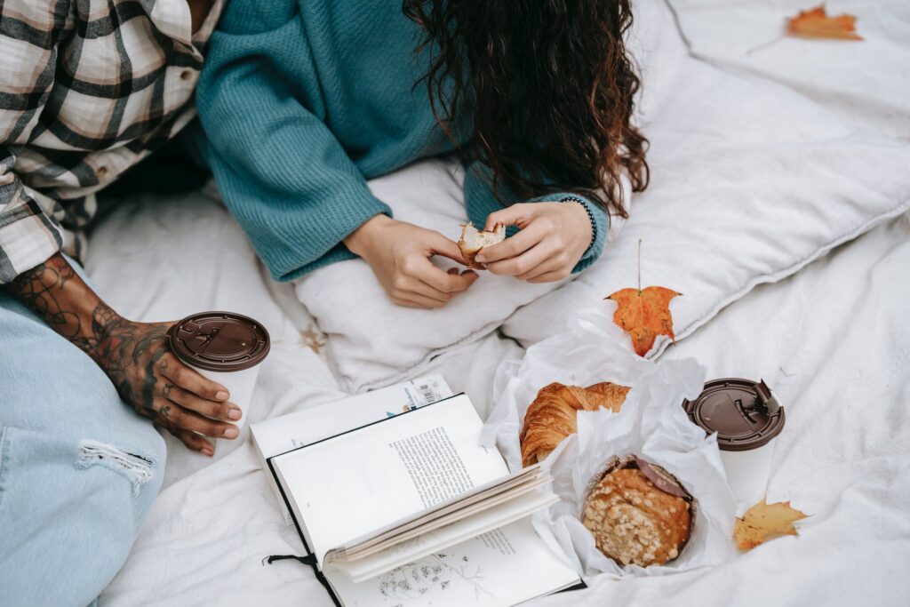 Stock photo from pexels by 
Tim Samuel of two people on a bed that has white bed sheets and a book on it with some breakfast items and autumn leafs