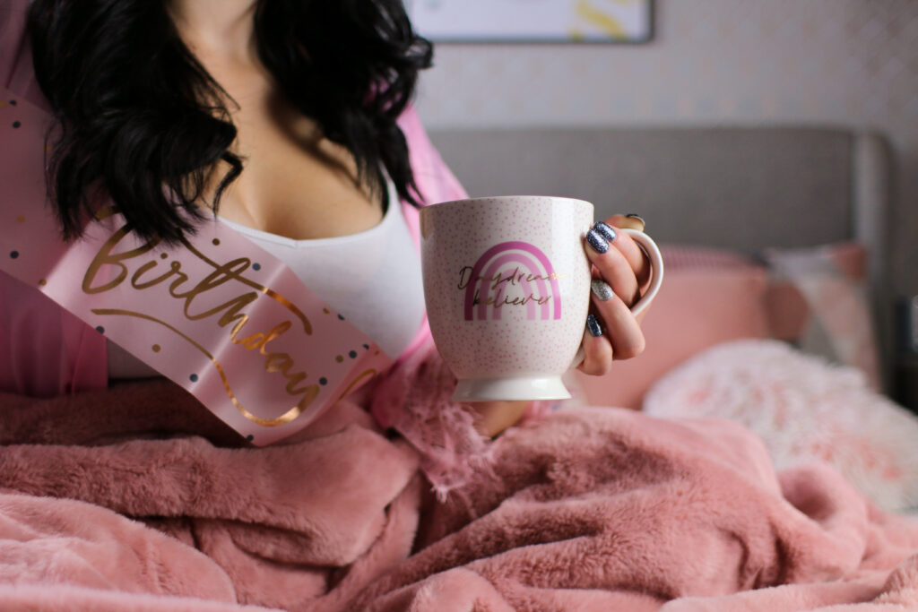 A close up photo of Jordanne sitting in a bed with a pink blanket wearing a pink robe and a pink birthday girl sash. She is holding a white mug that has "Daydream believer" written in gold.