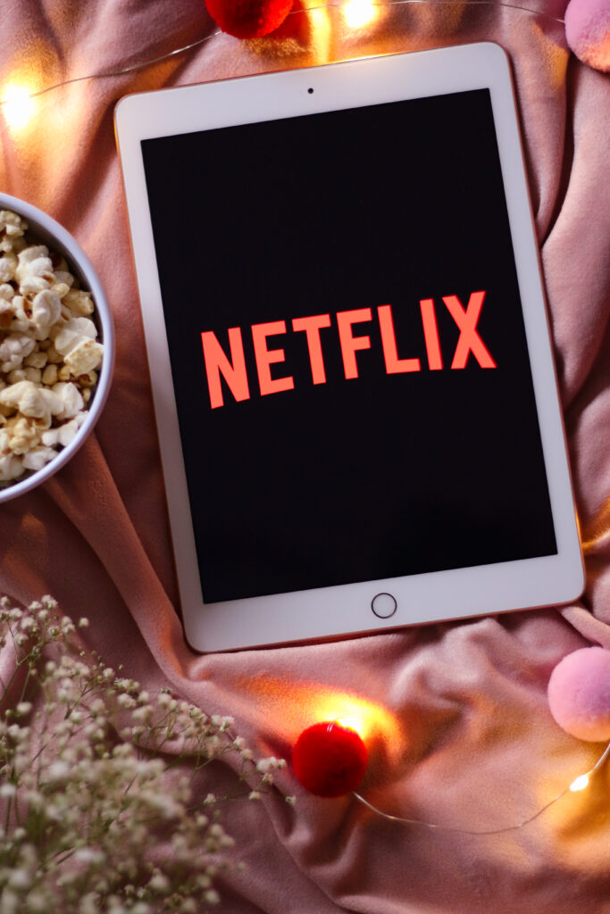 Birds eye view of an iPad with the Netflix logo on screen. A bowl of popcorn to the left and fairy lights around the iPad