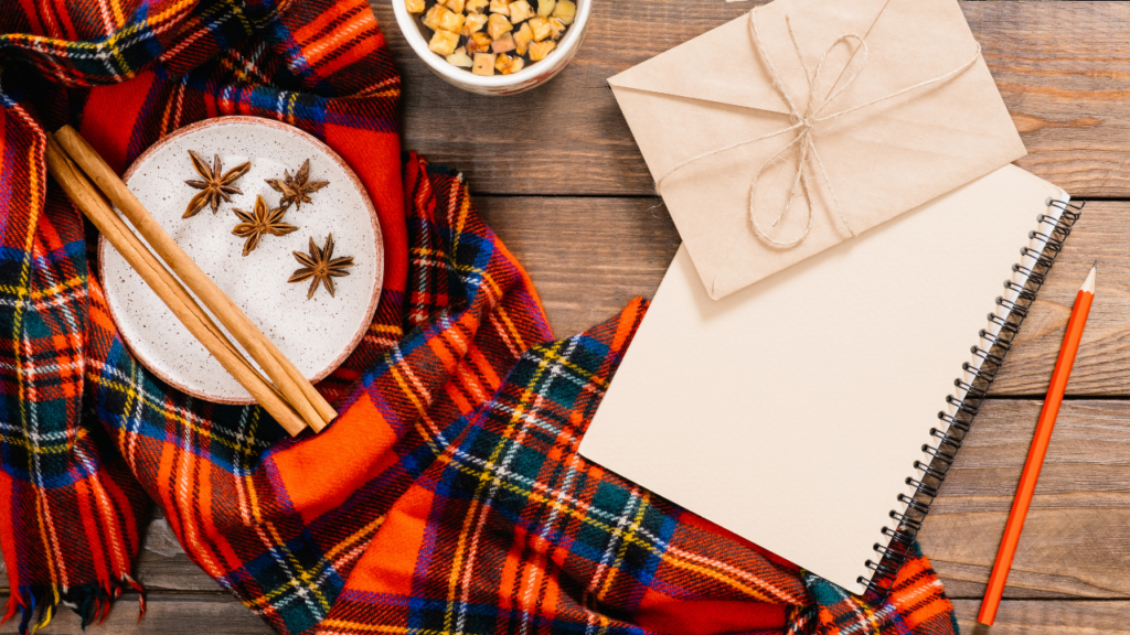 Stock photo from Canva of an autumn flatlay. There is a wooden work top in the background with a red tartan scarf in the left hand with a mug on it and a white notebook in the right hand of the photo