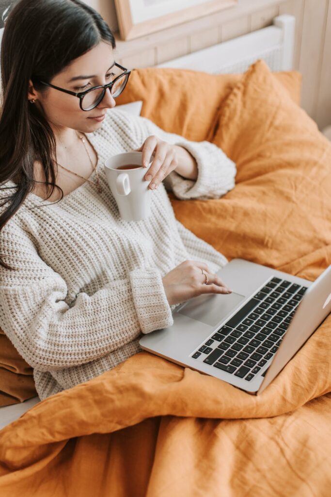 Stock photo from pexels by Vlada Karpovich of a woman with long brown hair and glasses sitting on a bed with mustard bedding. She is holding a white mug and looking at a laptop.