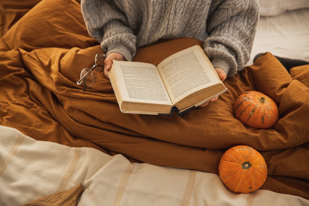 Stock photo from pexels by Monstera of a person sitting on a bed with a dark brown cover, an open book in hand and some orange pumpkins on the bed