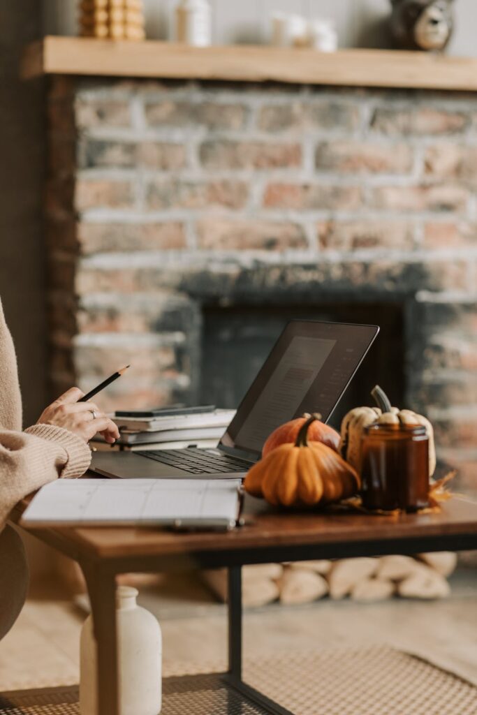 Stock photo from Pexels by Vlada Karpovich of a wooden table with a silver laptop on it with a brown candle and an orange pumpkin