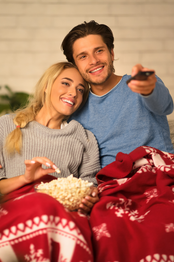 Mega list of Netflix UK Christmas Movies and Shows - Stock photo from Canva of a man and woman sitting on a couch with a red blanket and a bowl of popcorn watching tv