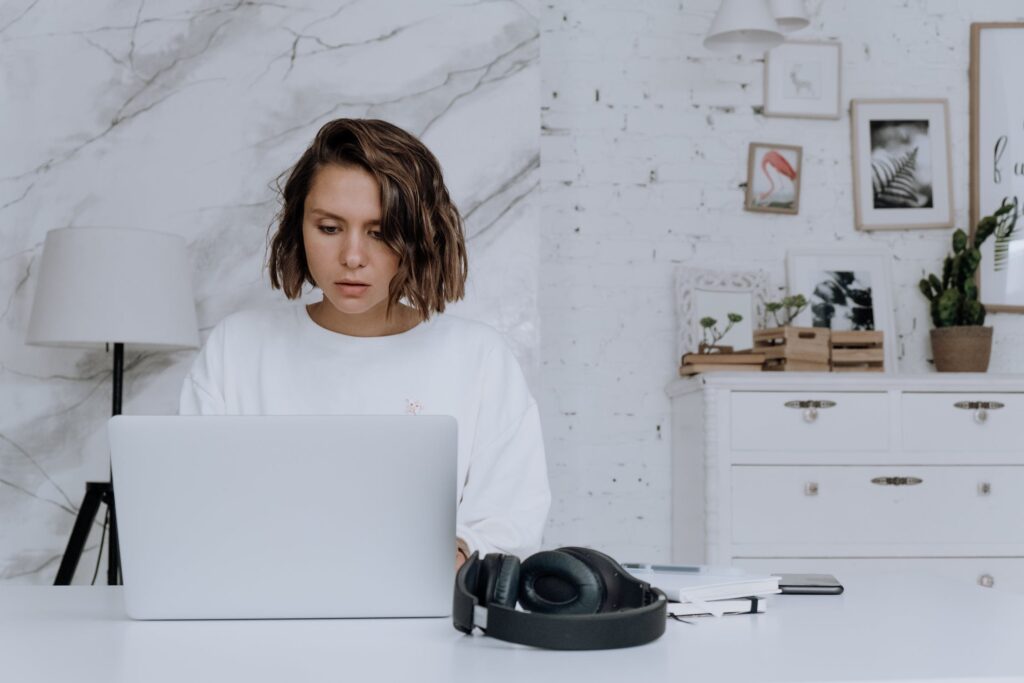 Stock photo from pexels by cottonbro of a woman with short brown hair wearing a white jumper sitting at a white desk typing on a silver laptop