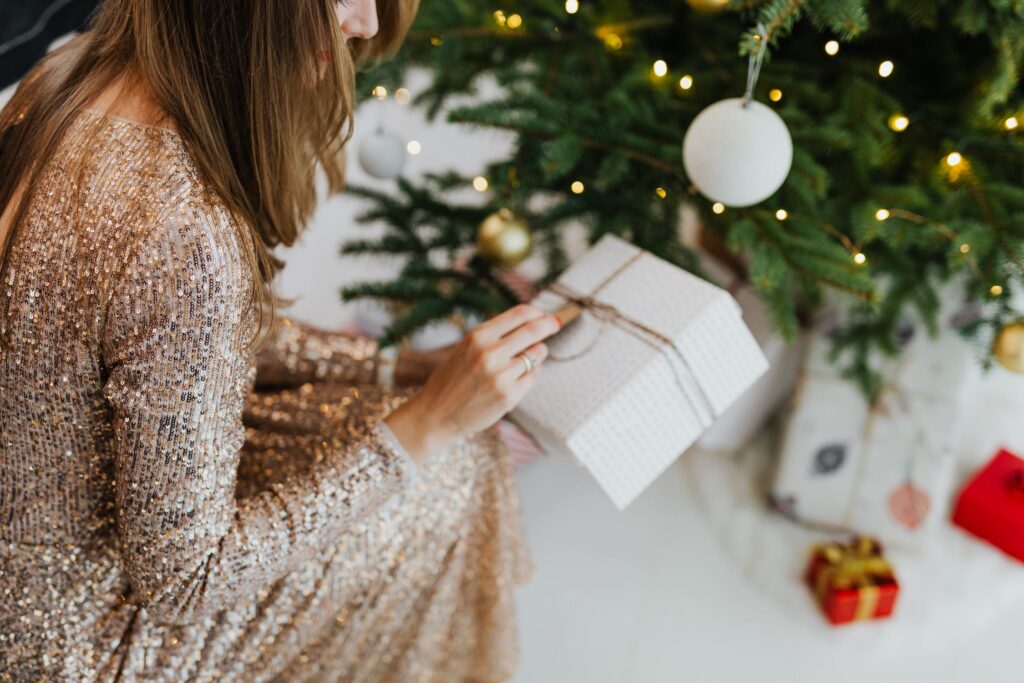 Stock photo from pexels by Karolina Grabowska of a woman wearing a gold sparkle long sleeve dress kneeling in front of a green christmas tree holding a white gift box with gold ribbon 