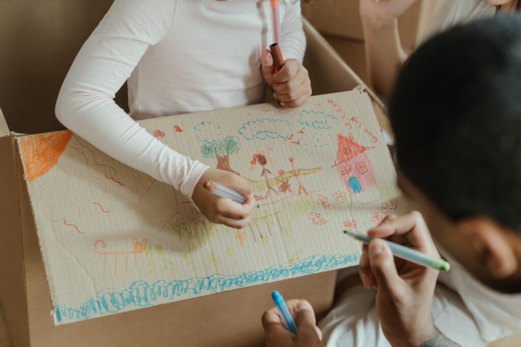 Stock photo from pexels by Cottonbro of a child in a cardboard box drawing on the box with colourful pens