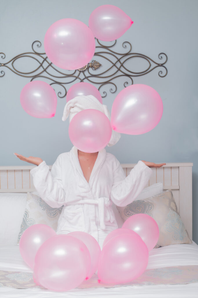 Stock photo from pexels by Dazzle Jam of a woman in white robe on a white bed with pink balloons all around her