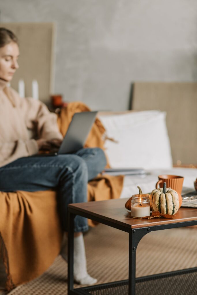 Stock photos from Pexels by Vlada Karpovich of woman sitting on cough with laptop and a brown table with candles and pumpkins in foreground