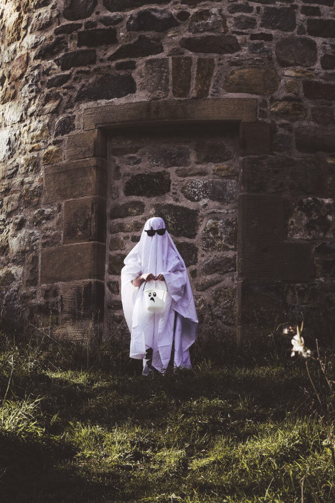 Photo of child wearing a white sheet and sunglasses to look like a ghost whilst holding a white bucket in woods
