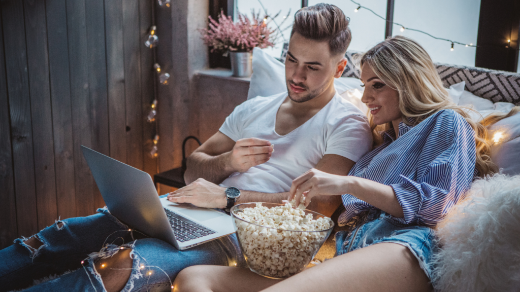 Stock photo on Canva by Svetikid at Getty Images of a woman and man on a bed watching movies with a bowl of popcorn