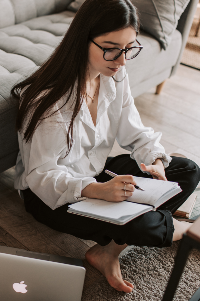 Stock photo from Pexels by Vlada Karpovich of a woman with long black hair and glasses writing in a white notebook
