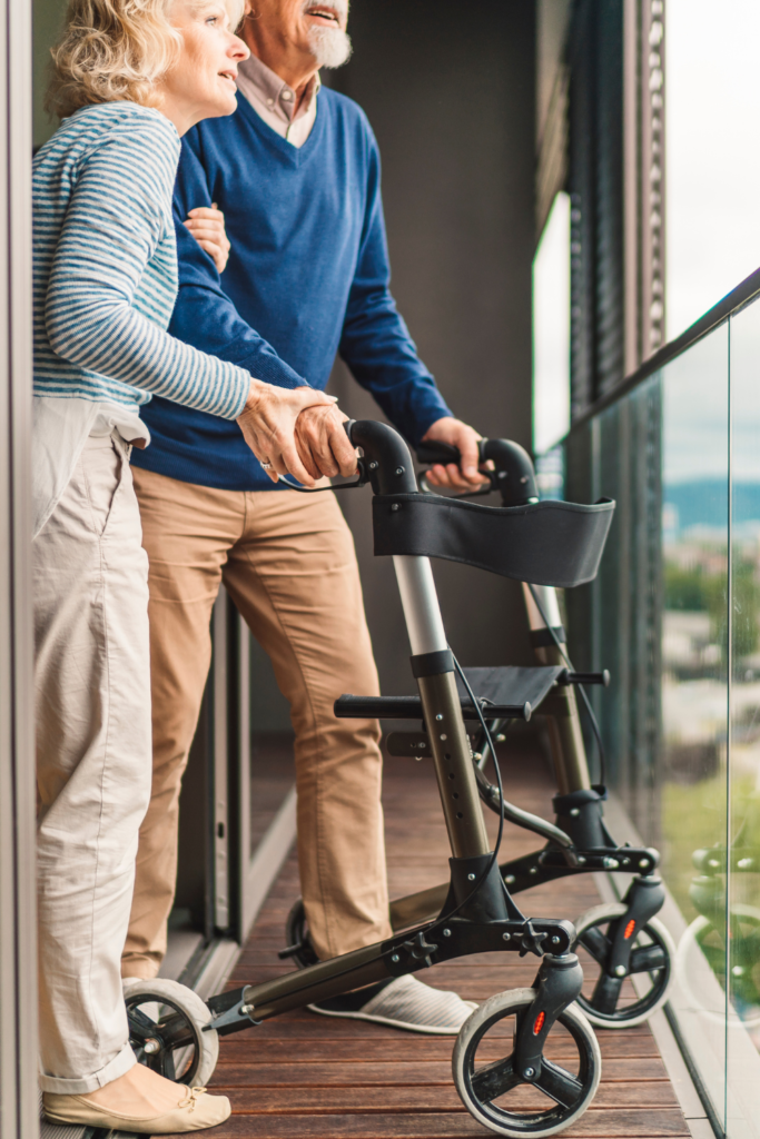 Stock photo from Canva by Ziga Plahutar of an elderly couple standing on a balcony looking out at a landscape whilst holding onto a Rollator Mobility Aid