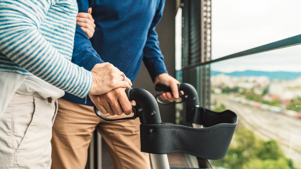 Stock photo from Canva by Ziga Plahutar of an elderly couple standing on a balcony looking out at a landscape whilst holding onto a Rollator
