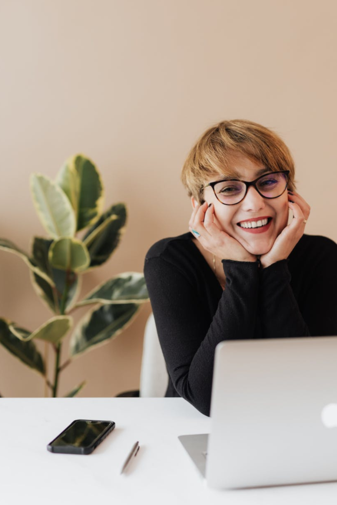 Stock photo from Pexels by Karolina Grabowska of a woman wearing glasses with short light brown hair smiling in front of a Laptop