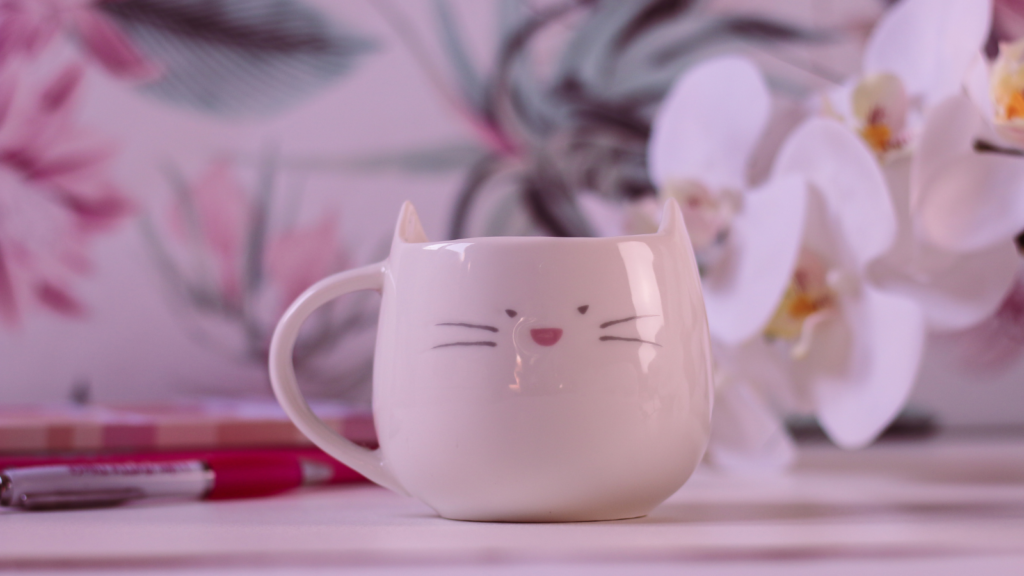 Close up photo of a white mug with a cat face. The mug is sitting on top of a white desk with a pink journal to the left hand side