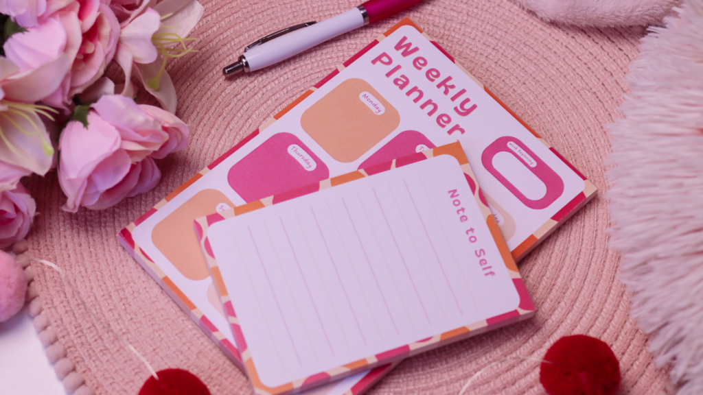 Birdseye view of a pink and orange weekly notepad planner and notes notepad by jordanneleecreative on a pink place mat with pink flowers to the left