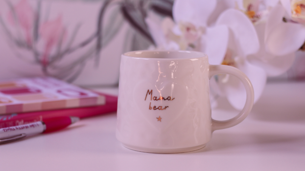 Close up photo of a white mug with gold text on it's side that reads "mama bear". The mug is sitting on top of a white desk with a pink journal to the left hand side