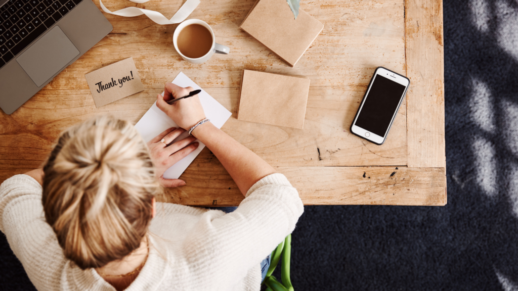 Stock photo from Canva by Monkey Business Images of a woman sat at a wooden desk writing in a journal