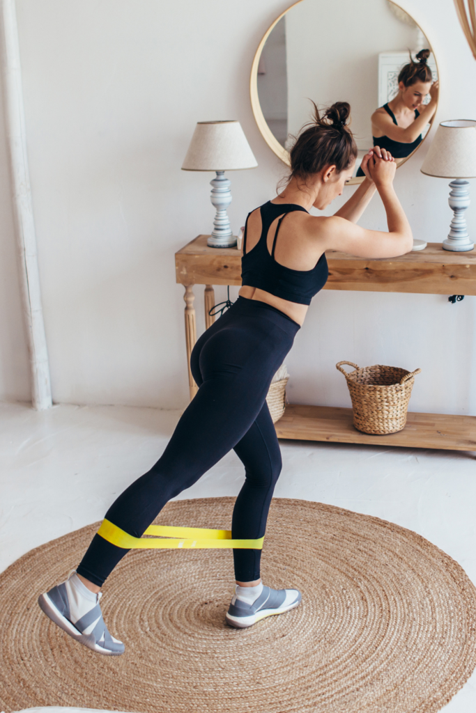 Stock photo from Canva by Undreys of a woman wearing black gym clothing and working out at home with a yellow resistance band on her ankles