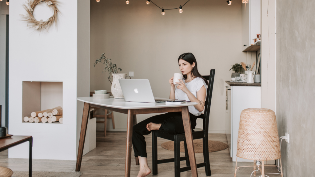 Stock photo from Canva by Vlada Karpovich of a woman sitting at a dining room table working on a silver apple macbook. The woman is holding a white mug