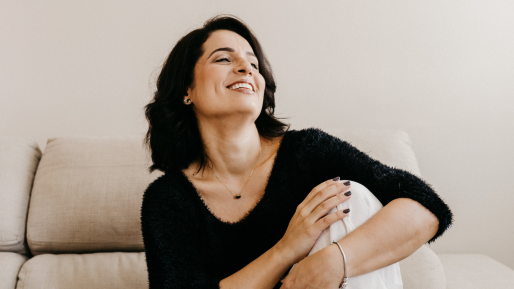 A stock photo from Canva of a woman wearing a black top and white pans sitting on a sofa smiling
