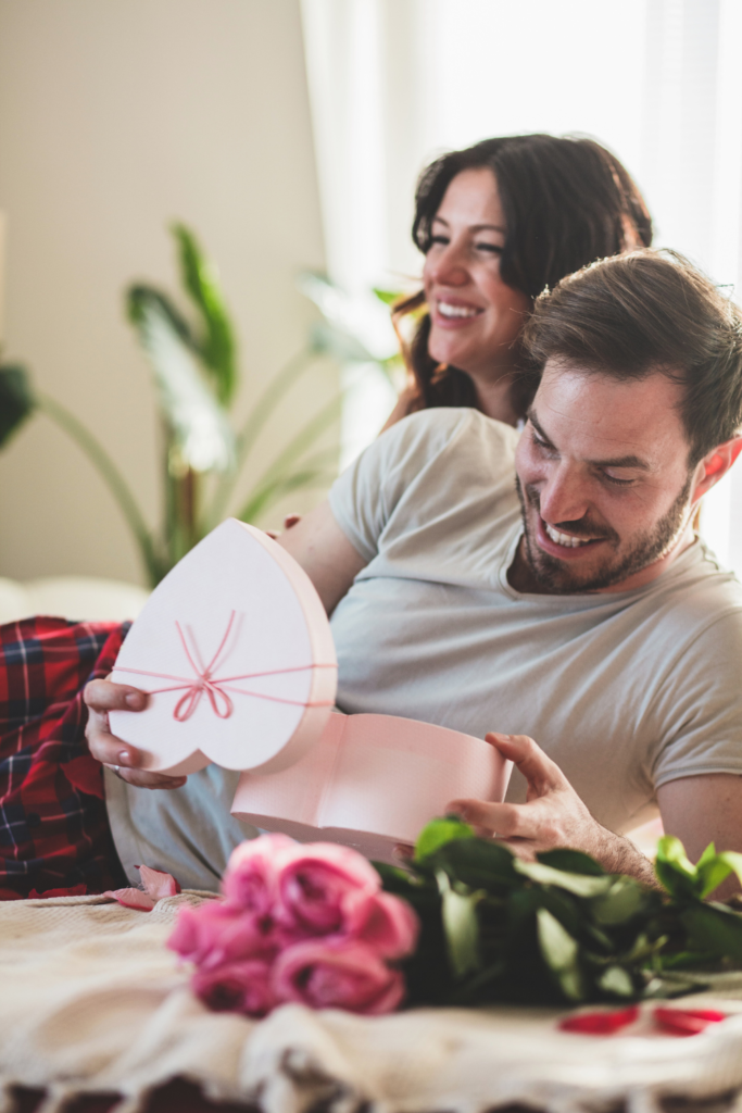 Stock photo from Canva of a couple laughing on a bed with a pink heart shaped box